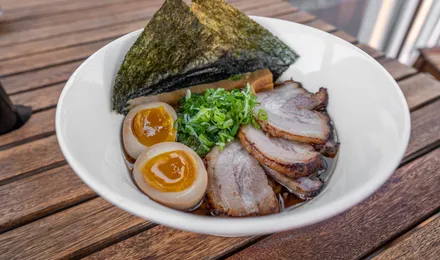 Ramen shoyu dans un bol large : nouilles, tranches de chashu, demi-œuf mariné au jaune coulant, nori et oignon vert dans un bouillon ambré.