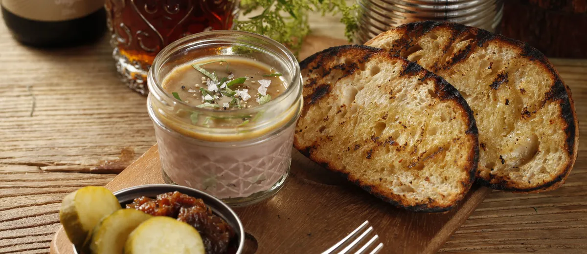 Rillettes du Mans dans un pot en grès, chair de porc effilochée en gros filaments dorés sous une couche de graisse, servies avec du pain de campagne et des cornichons