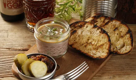 Rillettes du Mans dans un pot en grès, chair de porc effilochée en gros filaments dorés sous une couche de graisse, servies avec du pain de campagne et des cornichons